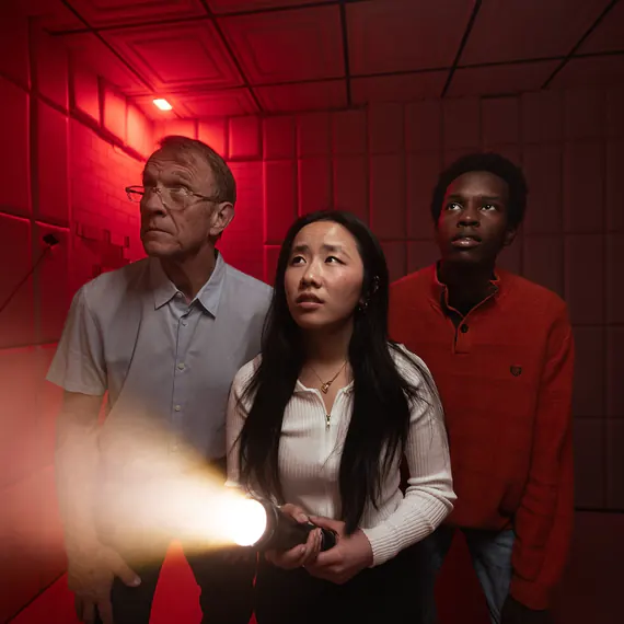 Three players in a red-lit cell, one holding a flashlight